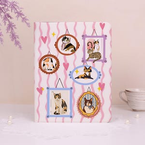 May include: A white notebook with a pink and white striped background. The notebook cover features framed illustrations of cats in various poses, with pink hearts and yellow stars scattered around the frames. The notebook is decorated with a lace trim.