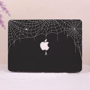 May include: Black laptop case featuring a white spiderweb design with spiders. The Apple logo is visible in white, with a spider hanging below it. The case is designed to fit a laptop.