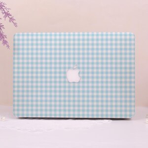 May include: A laptop with a light blue and white gingham pattern cover. The Apple logo is centered on the cover. The laptop is on a white surface.