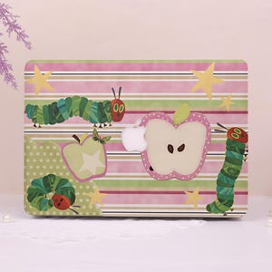 May include: A laptop hard shell case featuring a horizontal striped pattern in shades of pink, green, and white. The design includes illustrations of green caterpillars, yellow stars, and pink and light green apples. A central cutout reveals an Apple logo.