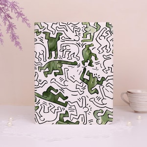 Pode incluir: Um caderno retangular com fundo branco e um padrão repetido de figuras verdes em várias poses, delineadas em preto. O caderno está sobre uma superfície branca. O design lembra a arte de Keith Haring.