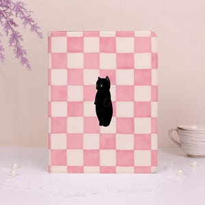 May include: A pink and white checkered notebook with a black cat illustration. The cat has yellow eyes and is standing upright. The notebook is rectangular and appears to be closed.
