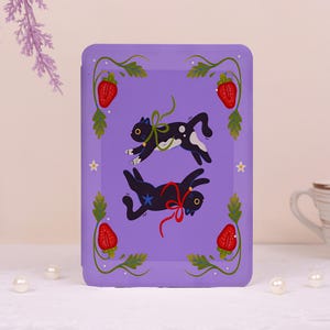 May include: A rectangular purple e-reader case featuring two black cats leaping in the center. One cat wears a green ribbon, the other a red ribbon. The border is decorated with green vines, red strawberries, and small white flowers.