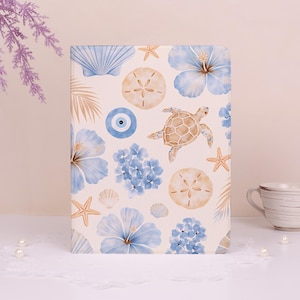 Può includere: Un taccuino con sfondo color crema, decorato con un design ad acquerello di creature marine e fiori. Il design include fiori di ibisco blu, stelle marine, conchiglie, una tartaruga e un simbolo dell'occhio blu.