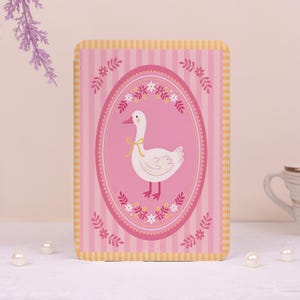 May include: A rectangular card featuring a pink background with vertical stripes and a yellow checkered border. The center displays an oval frame containing a white goose with a yellow ribbon around its neck, surrounded by small floral motifs.
