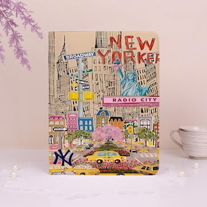 Op de afbeelding: Een notitieboek met een kleurrijke illustratie van New York City. De omslag toont iconische bezienswaardigheden zoals het Vrijheidsbeeld, Broadway-borden en gele taxi's. De woorden "New Yorker" en "Radio City" zijn ook zichtbaar.