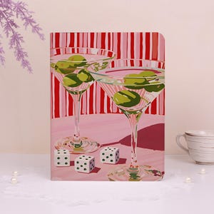 May include: A rectangular notebook cover featuring a vibrant illustration of two martini glasses filled with green olives, set against a background of vertical red and white stripes. Three white dice with black dots are positioned in the foreground.