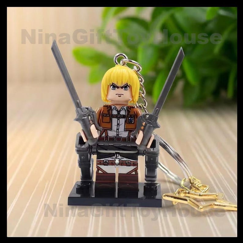 K&ouml;nnte beinhalten: Ein Schl&uuml;sselanh&auml;nger mit einer blonden Anime-Minifigur in einer braun-wei&szlig;en Uniform mit Geschirr. Die Figur h&auml;lt zwei graue Schwerter in aufrechter Position und steht auf einem schwarzen Sockel. Der Text NinaGiftToyHouse ist sichtbar.
