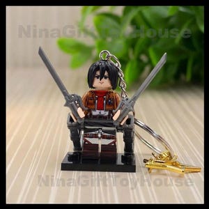 K&ouml;nnte beinhalten: Eine Miniatur-Schl&uuml;sselanh&auml;nger-Figur mit schwarzem Haar, rotem Schal und brauner Jacke &uuml;ber einem wei&szlig;en Hemd. Die Figur h&auml;lt zwei graue Schwerter in den H&auml;nden und ist an einem silbernen Metallschl&uuml;sselring mit einem kleinen goldenen Anh&auml;nger befestigt.