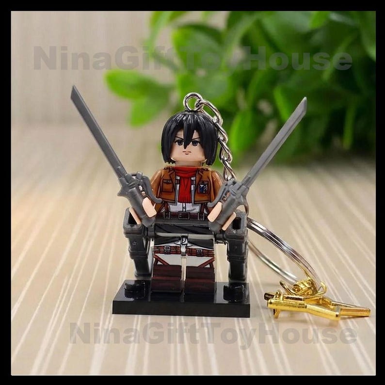 K&ouml;nnte beinhalten: Eine Miniatur-Schl&uuml;sselanh&auml;nger-Figur mit schwarzem Haar, rotem Schal und brauner Jacke &uuml;ber einem wei&szlig;en Hemd. Die Figur h&auml;lt zwei graue Schwerter in den H&auml;nden und ist an einem silbernen Metallschl&uuml;sselring mit einem kleinen goldenen Anh&auml;nger befestigt.