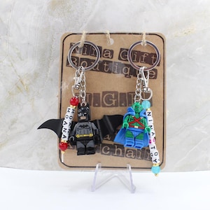 May include: Two keychains featuring plastic superhero figures attached to metal rings and chains. One keychain has a black Batman figure with wings and red beads spelling MOANA. The other has a green Martian Manhunter figure with blue beads spelling NINA TOY.