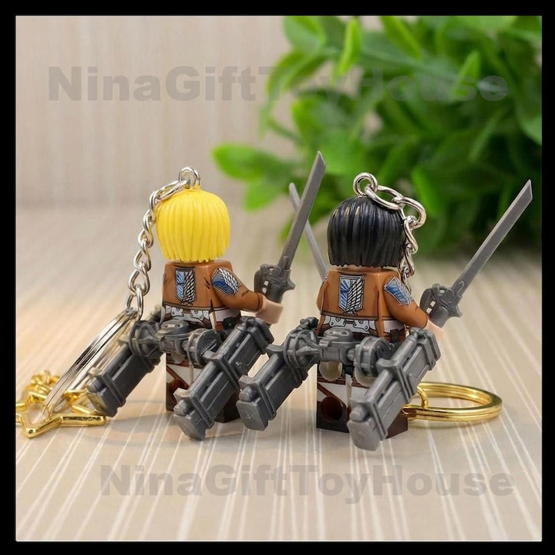K&ouml;nnte beinhalten: Zwei Figuren-Schl&uuml;sselanh&auml;nger mit gelbem und schwarzem Haar, die braune Jacken mit blau-wei&szlig;en Fl&uuml;gel-Emblemen auf dem R&uuml;cken tragen. Jede Figur ist mit grauer mechanischer Ausr&uuml;stung und Schwertern an einer silbernen Metallkette ausgestattet.