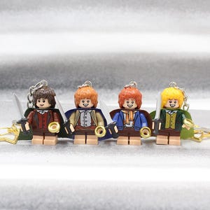 May include: Four miniature plastic character keychains featuring figures with brown, orange, and yellow hair. Each figure wears a colorful cape, a detailed vest, and holds a small silver-colored sword in one hand and a gold-colored ring in the other.