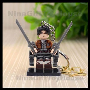 K&ouml;nnte beinhalten: Ein Miniatur-Schl&uuml;sselanh&auml;nger aus Kunststoff, der eine Figur mit schwarzen Haaren zeigt, die eine braune Jacke &uuml;ber einem grauen Hemd und eine dunkle Hose tr&auml;gt. Die Figur h&auml;lt zwei graue Schwerter und ist an einer silbernen Metallkette befestigt.
