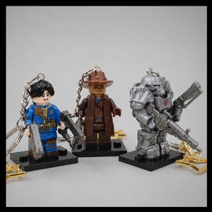 May include: Three character keychains featuring plastic figures on black bases. One wears a blue jumpsuit, one is a brown-clad figure with a hat, and one is a silver-colored armored figure holding a weapon. Each has a metal chain attached.