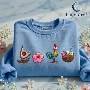 a blue t-shirt with various colorful and whimsical embroidered designs, including a sailboat, a flower, a coconut, and a rooster, all set against a marble background with flowers.
