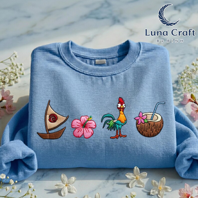 a blue t-shirt with various colorful and whimsical embroidered designs, including a sailboat, a flower, a coconut, and a rooster, all set against a marble background with flowers.