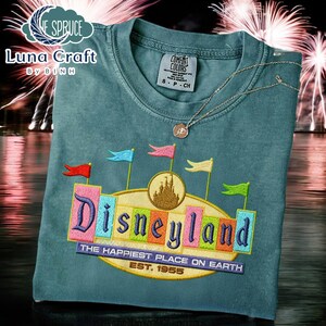 A t-shirt with a colorful embroidered design of the Disneyland logo, featuring a castle and flags, against a background of fireworks.