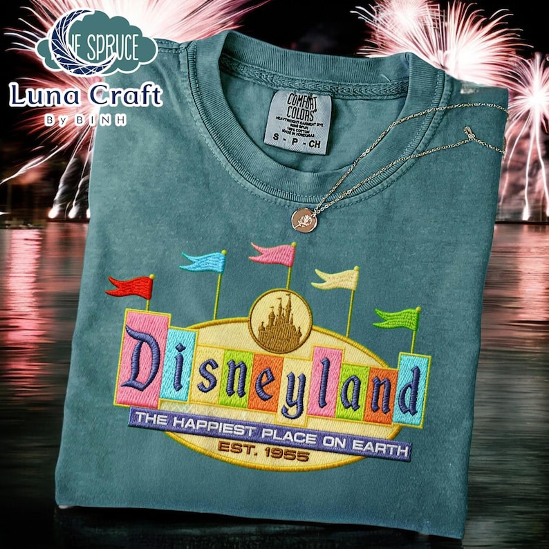 A t-shirt with a colorful embroidered design of the Disneyland logo, featuring a castle and flags, against a background of fireworks.