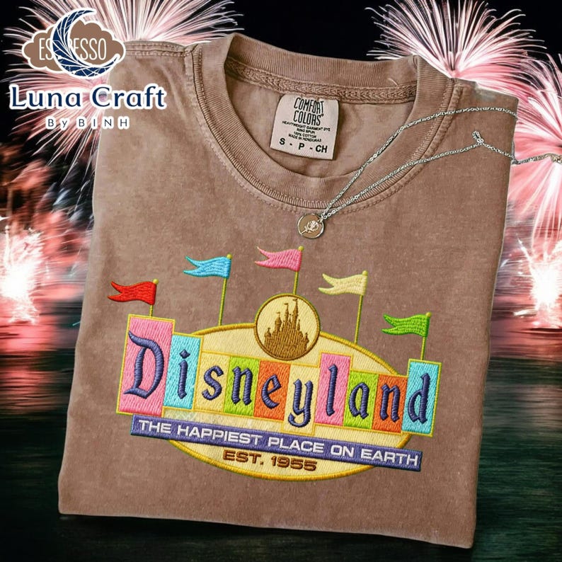 A brown t-shirt with a colorful embroidered design of Disneyland, featuring a castle and flags, is displayed against a background of fireworks.