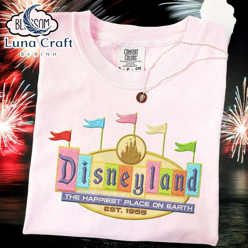 A pink t-shirt with a colorful embroidered design of Disneyland, featuring a castle and flags, is displayed against a background of fireworks.