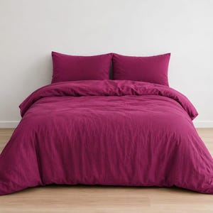 May include: A solid magenta bedding set consisting of a duvet cover and two matching rectangular pillowcases. The fabric has a matte finish and a slightly textured appearance, draped neatly over a bed frame to create a uniform color display.