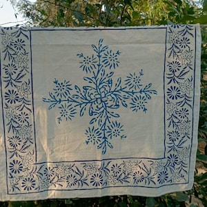 Puede incluir: Textil blanquecino con un diseño floral azul. El motivo central es un arreglo floral estilizado, rodeado por un borde de patrones florales repetidos. El textil está colgado al aire libre, con vegetación de fondo.