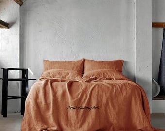 May include: A bed dressed with terracotta-coloured linen bedding, including a duvet cover and pillowcases. The bedding is draped over the bed, creating soft folds. The room has a minimalist design with a white wall and a black side table.