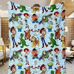 May include: A light blue blanket featuring a repeating pattern of characters from the animated film Toy Story. The design includes Woody, Buzz Lightyear, Jessie, Mr. Potato Head, Rex, Slinky Dog, and other characters. The blanket has a cloud-filled sky background.
