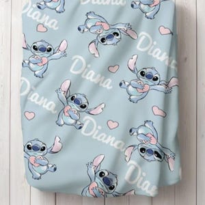May include: A light blue blanket featuring a repeating pattern of the blue alien character Stitch holding a pink heart, interspersed with small pink hearts and the name Diana written in a white cursive script across the fabric surface.