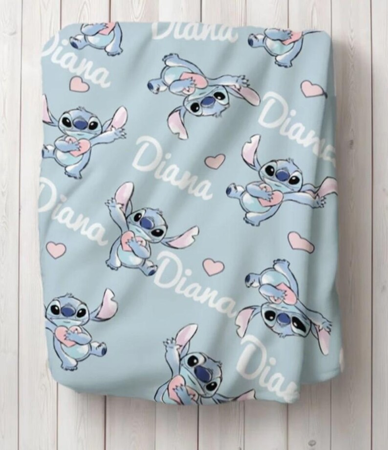 May include: A light blue blanket featuring a repeating pattern of the blue alien character Stitch holding a pink heart, interspersed with small pink hearts and the name Diana written in a white cursive script across the fabric surface.