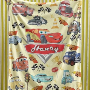 Peut inclure: Une couverture cr&egrave;me avec des voitures de dessins anim&eacute;s, des drapeaux &agrave; damiers et le nom "Henry" en rouge. La couverture pr&eacute;sente diverses illustrations de voitures, dont une voiture de course rouge, un camion de pompiers et une voiture de police.