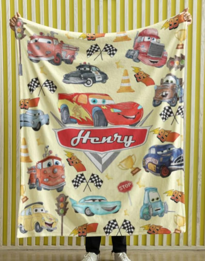 Peut inclure: Une couverture cr&egrave;me avec des voitures de dessins anim&eacute;s, des drapeaux &agrave; damiers et le nom "Henry" en rouge. La couverture pr&eacute;sente diverses illustrations de voitures, dont une voiture de course rouge, un camion de pompiers et une voiture de police.