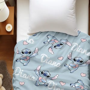 May include: A light blue blanket featuring a repeating pattern of the blue character Stitch holding a pink heart, interspersed with small pink hearts and the name Diana written in a white cursive script across the fabric surface.