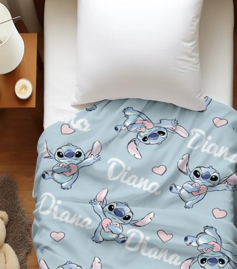 May include: A light blue blanket featuring a repeating pattern of the blue character Stitch holding a pink heart, interspersed with small pink hearts and the name Diana written in a white cursive script across the fabric surface.