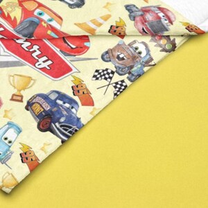 Peut inclure: Une couverture jaune avec un motif de voitures de course de dessins anim&eacute;s, de drapeaux &agrave; damier et de troph&eacute;es. Le nom "Henry" est &eacute;crit sur une enseigne rouge. La couverture a une bordure blanche et est pos&eacute;e sur un fond jaune.