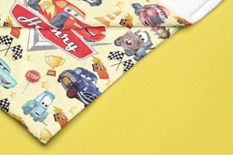 Peut inclure: Une couverture jaune avec un motif de voitures de course de dessins anim&eacute;s, de drapeaux &agrave; damier et de troph&eacute;es. Le nom "Henry" est &eacute;crit sur une enseigne rouge. La couverture a une bordure blanche et est pos&eacute;e sur un fond jaune.