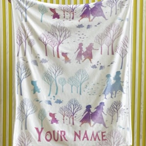 May include: White blanket featuring a winter forest scene with purple and blue trees and figures. The blanket has the text "YOUR NAME" in pink. The blanket is held up by a person.