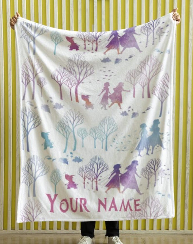 May include: White blanket featuring a winter forest scene with purple and blue trees and figures. The blanket has the text "YOUR NAME" in pink. The blanket is held up by a person.