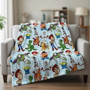 May include: A light blue blanket featuring a repeating pattern of characters from the Toy Story movie franchise. The blanket is draped over a light gray sofa. Characters include Woody, Buzz Lightyear, and others.