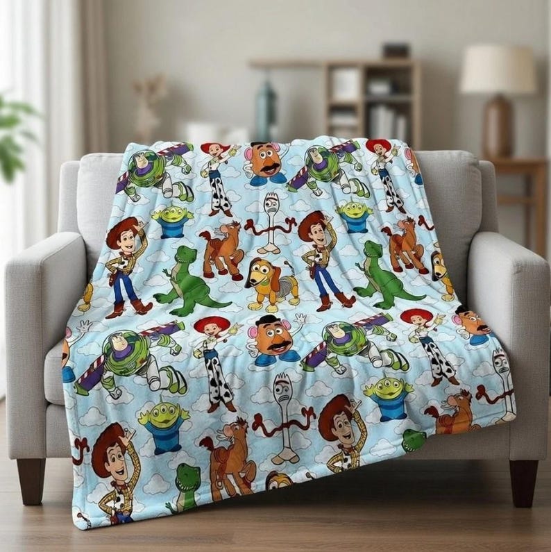 May include: A light blue blanket featuring a repeating pattern of characters from the Toy Story movie franchise. The blanket is draped over a light gray sofa. Characters include Woody, Buzz Lightyear, and others.
