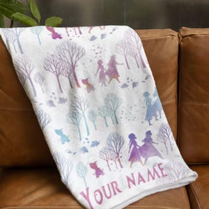 May include: White blanket with a repeating pattern of stylized trees and figures in purple, blue, and pink. The blanket is draped over a brown leather couch. The text "YOUR NAME" is printed on the blanket.