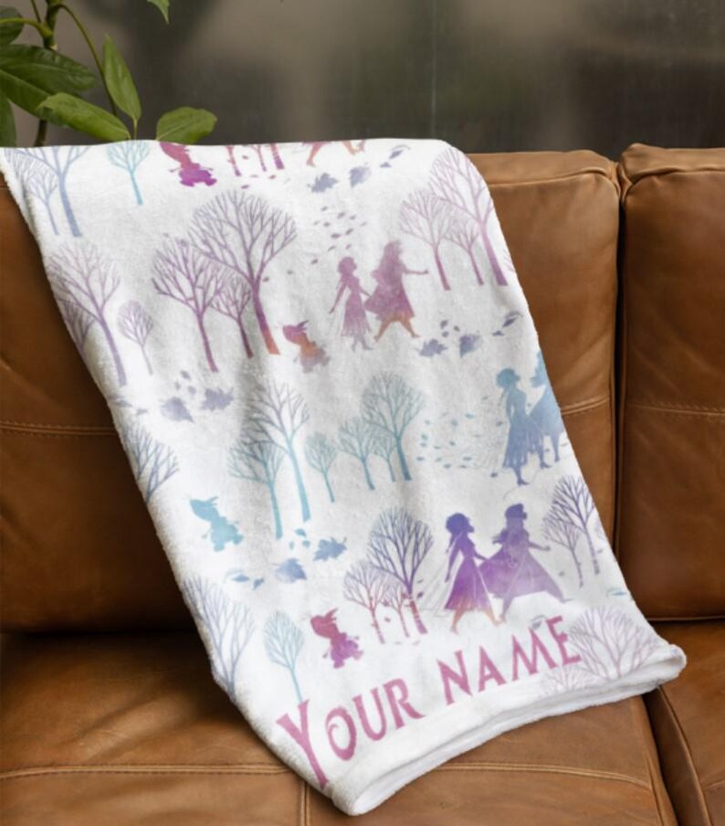 May include: White blanket with a repeating pattern of stylized trees and figures in purple, blue, and pink. The blanket is draped over a brown leather couch. The text "YOUR NAME" is printed on the blanket.