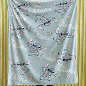 May include: A light blue fleece blanket featuring a repeating pattern of the character Stitch holding a pink heart, interspersed with small pink hearts and the name Diana written in a white cursive script across the fabric surface.