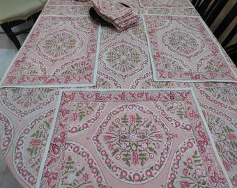 Op de afbeelding: Een roze bloemen tafelkleed met bijpassende placemats en servetten. Het tafelkleed en de placemats hebben een herhalend patroon van bloemen en bladeren in roze, groen en wit. De servetten zijn op een zwart bord gestapeld. Bananen liggen op tafel.