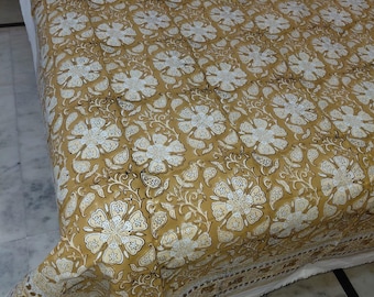 May include: A mustard-colored floral patterned bedspread with white flowers and leaves. The bedspread has a white border and is draped over a bed. The floral pattern is repeated across the fabric.