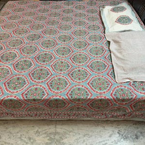 May include: A bedspread featuring a repeating geometric pattern of teal, red, and green floral motifs on a light blue background, accompanied by two rectangular pillows, one with a matching floral design and one with a fine striped pattern.