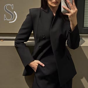 Puede incluir: Un blazer negro con diseño de doble botonadura y solapa con muescas. El blazer tiene dos botones visibles y un bolsillo. La imagen también muestra un pantalón negro a juego. El estilo general es profesional y a medida.