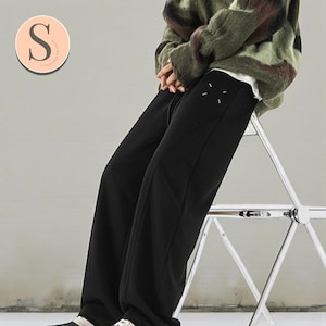 Könnte beinhalten: Schwarze Jogginghose mit weißem Nahtdesign am linken Bein. Die Hose wird mit einem grün-braunen Camouflage-Pullover kombiniert. Das Bild zeigt auch ein Logo mit dem Buchstaben "S" in einem Kreis.