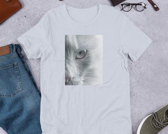 May include: White t-shirt featuring a close-up image of a white cat's face. The image is in grayscale, highlighting the cat's eye and fur. The shirt is laid flat on a textured surface, with accessories like a watch and wallet.